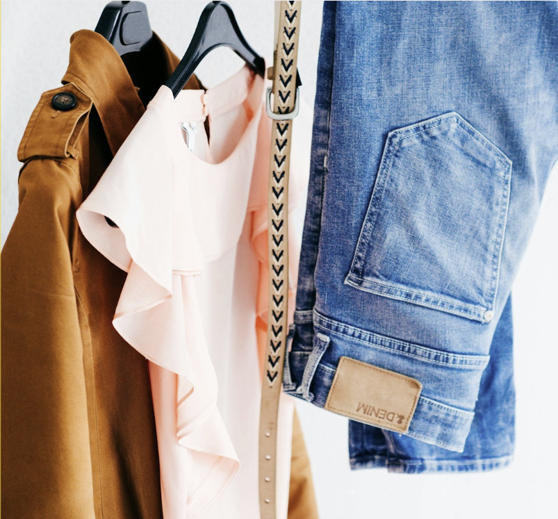 Styled clothing on hangers featuring a camel trench coat, pink ruffled blouse, patterned belt, and denim jeans
